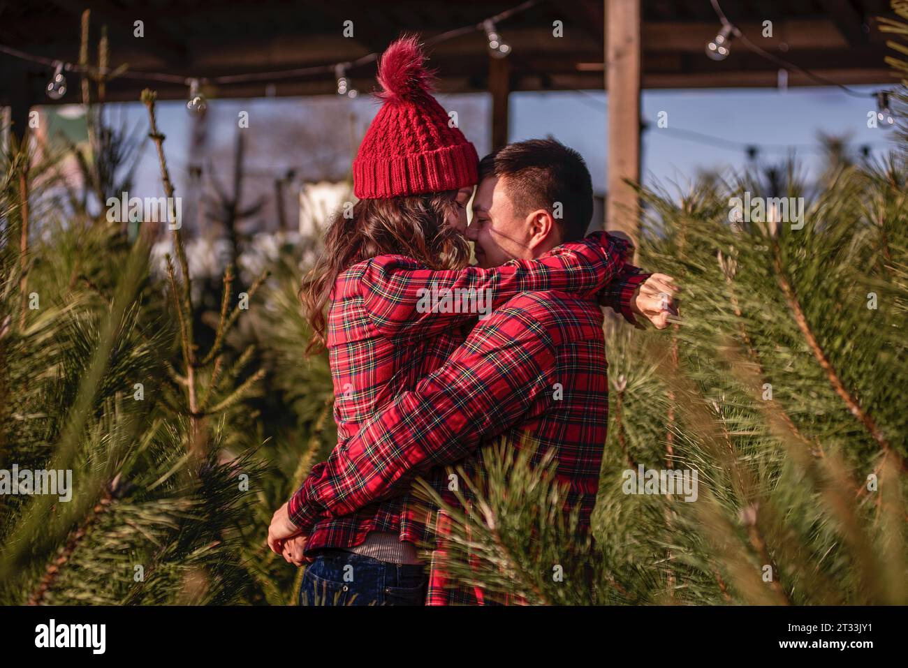 Bella coppia innamorata in camicie rosse a scacchi, cappelli a maglia si abbracciano strettamente naso a naso faccia a faccia tra i verdi mercatini dell'albero di Natale. Il giovane laug Foto Stock