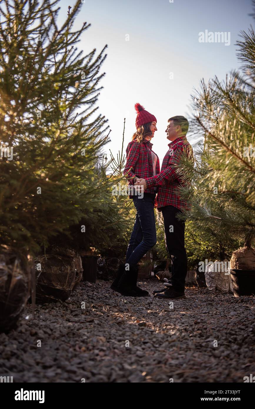 Coppia allegra innamorata in camicie rosse a scacchi, cappelli a maglia che si tengono per mano tra il verde mercato dell'albero di Natale. Il giovane guarda con amore la bella donna Foto Stock