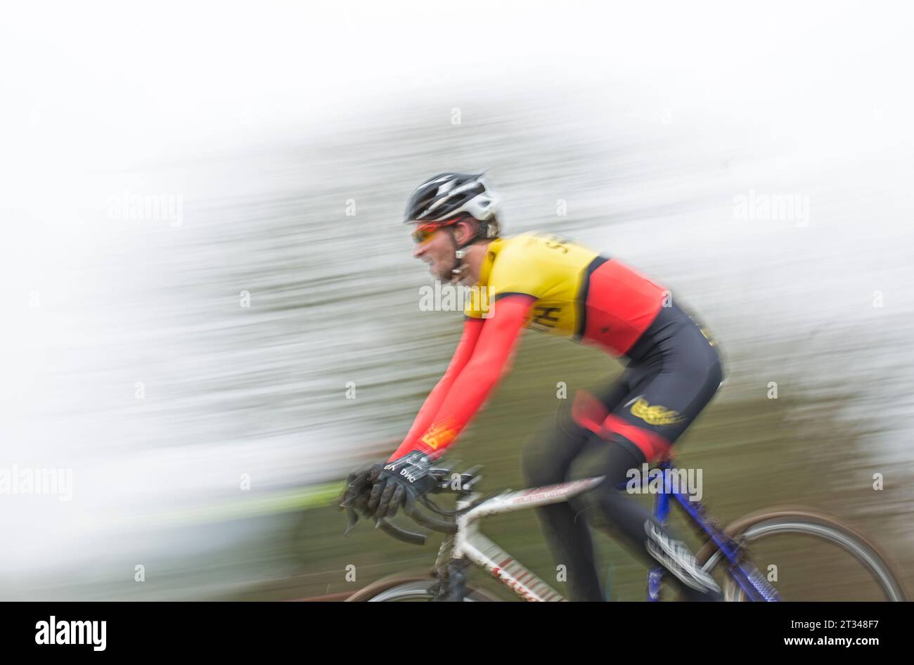 Immagine sfocata/in movimento di un pilota ciclocross maschile che corre in velocità attraverso una fila o alberi in una gara. Foto Stock