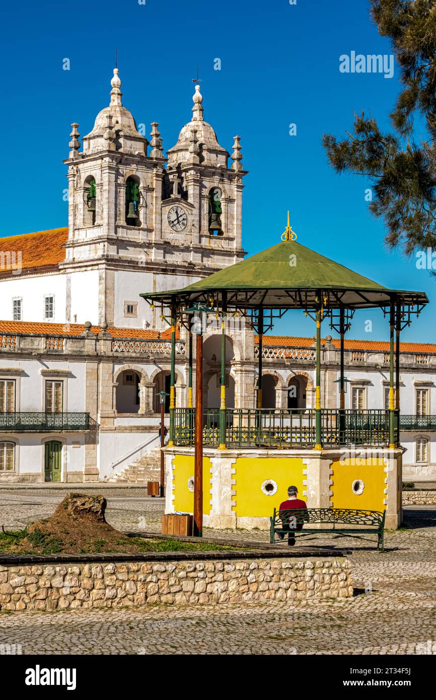 Nazaré, Portogallo - febbraio 2023: Piazza della città vecchia di Nazaré con una tribuna gialla e un uomo seduto su una panchina di fronte alla chiesa di Nossa Senhora da Nazaré. Foto Stock