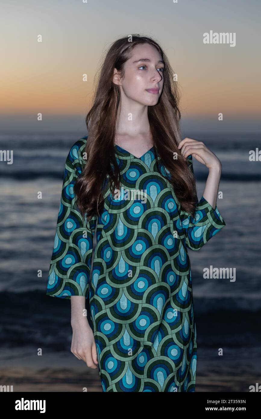 Una donna caucasica di 20 anni si erge sulla spiaggia al tramonto, indossando un vivace e colorato abito a camicia mentre il sole emana un caldo bagliore Foto Stock