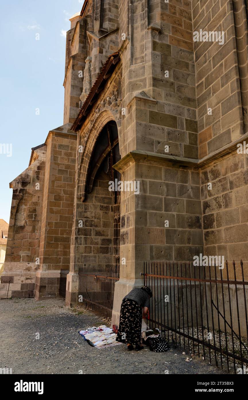 Anziana donna del posto che organizza i suoi beni in vendita all'esterno dell'iconica chiesa nera di Brasov. Foto Stock