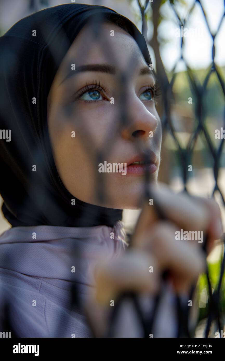 Donna che indossa hijab che contempla dietro una recinzione di chainlink Foto Stock