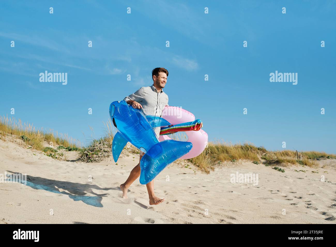 Uomo felice che corre con l'anello gonfiabile e lo squalo giocattolo in spiaggia nelle giornate di sole Foto Stock