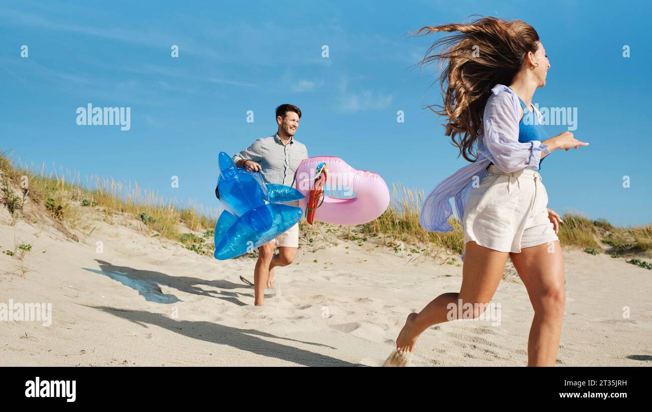 Donna felice che corre con un uomo che tiene l'anello gonfiabile e lo squalo giocattolo Foto Stock