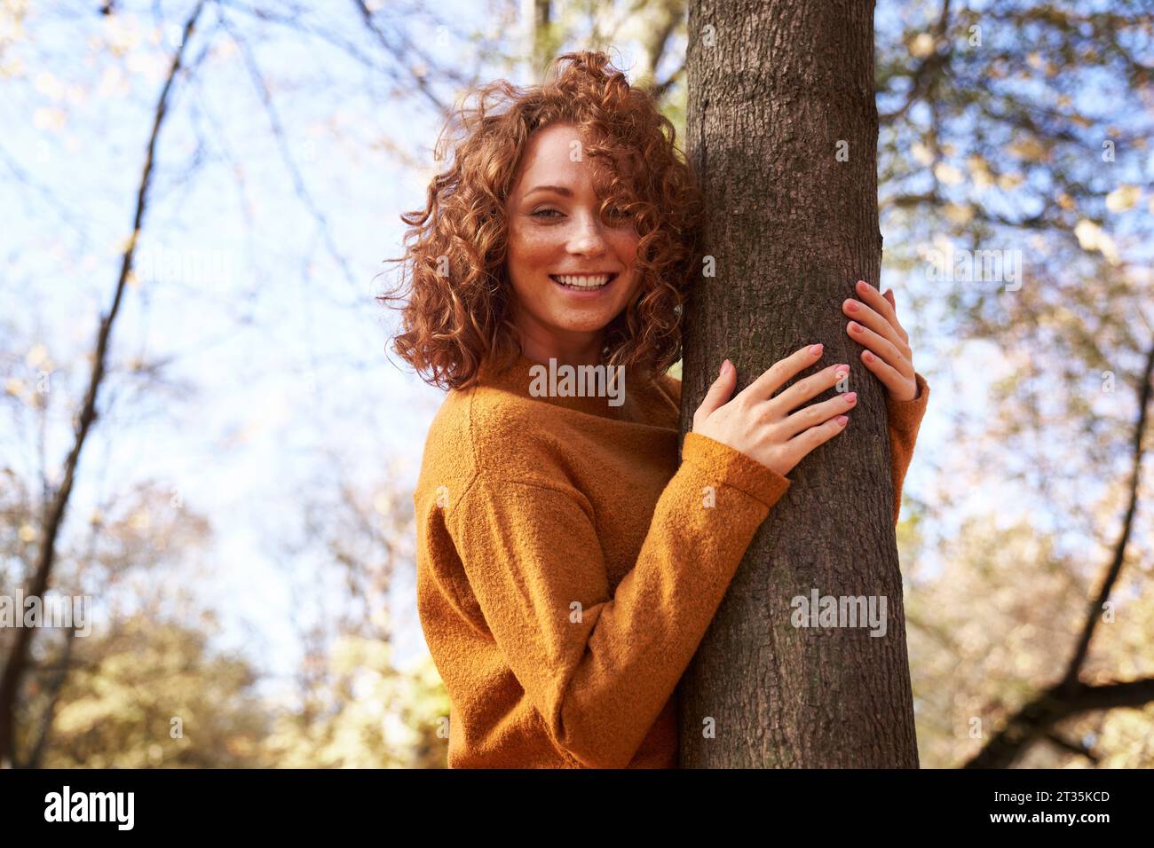 Donna rossa sorridente che abbraccia l'albero al parco Foto Stock