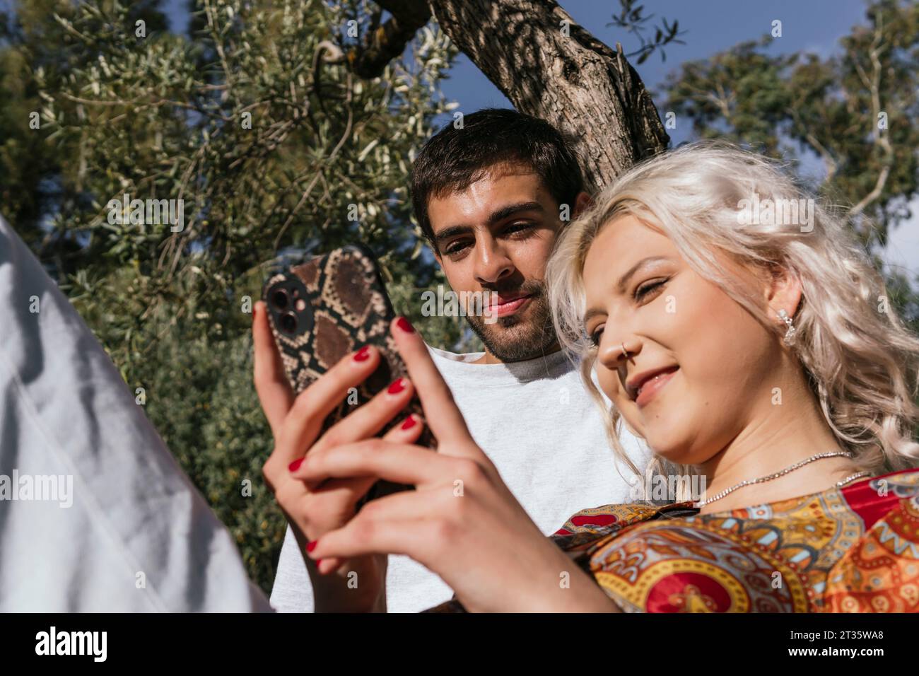 Fidanzata sorridente che mostra e usa lo smartphone con fidanzato davanti all'albero Foto Stock