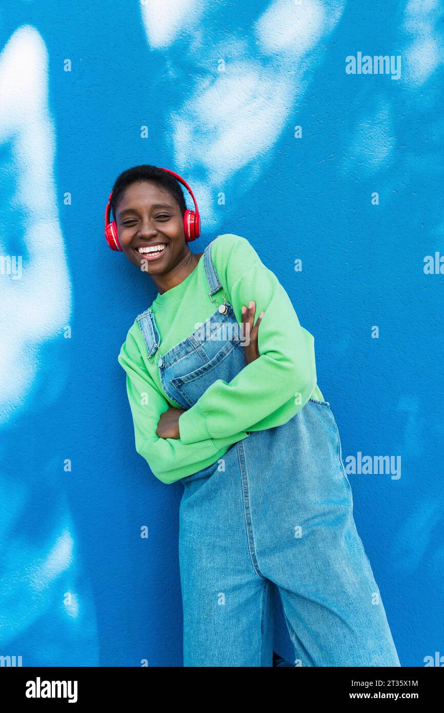 Donna felice che indossa cuffie wireless in piedi con le braccia incrociate davanti alla parete blu Foto Stock