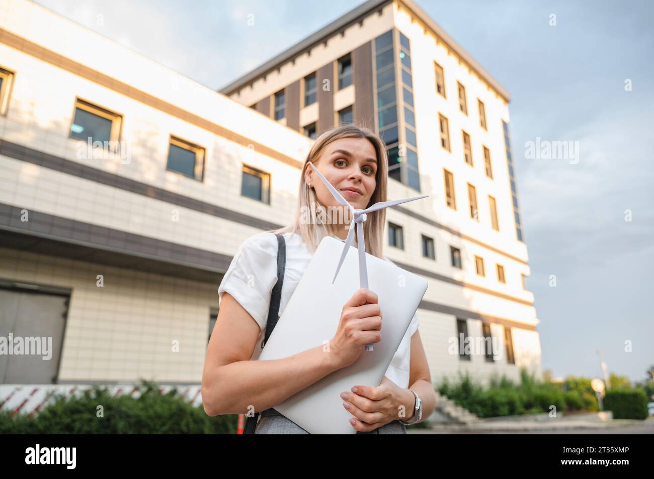 Donna d'affari sorridente in piedi con un modello di turbina eolica di fronte all'edificio Foto Stock