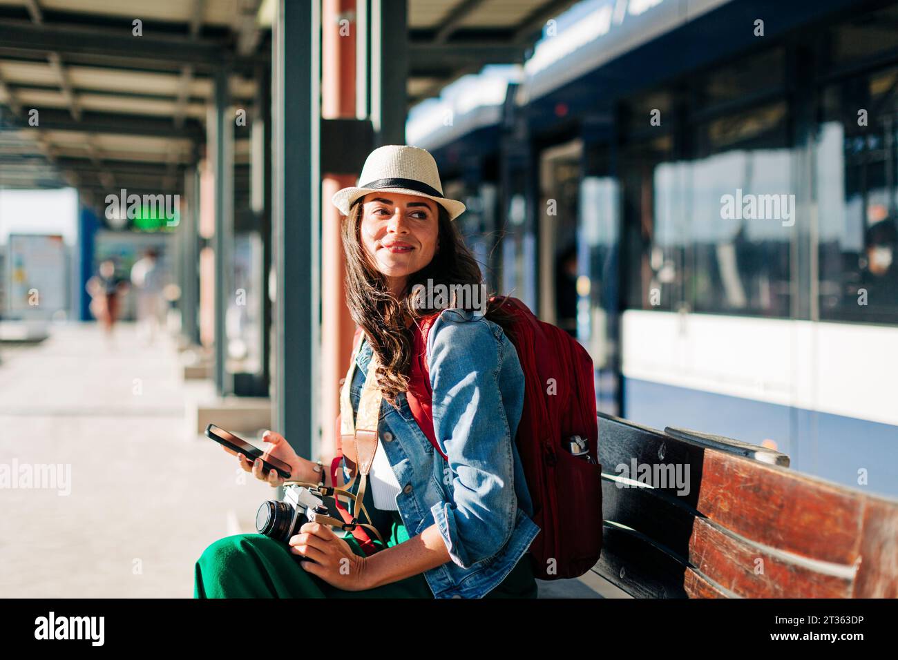 Donna felice che indossa un cappello seduto sulla panchina alla stazione del tram Foto Stock