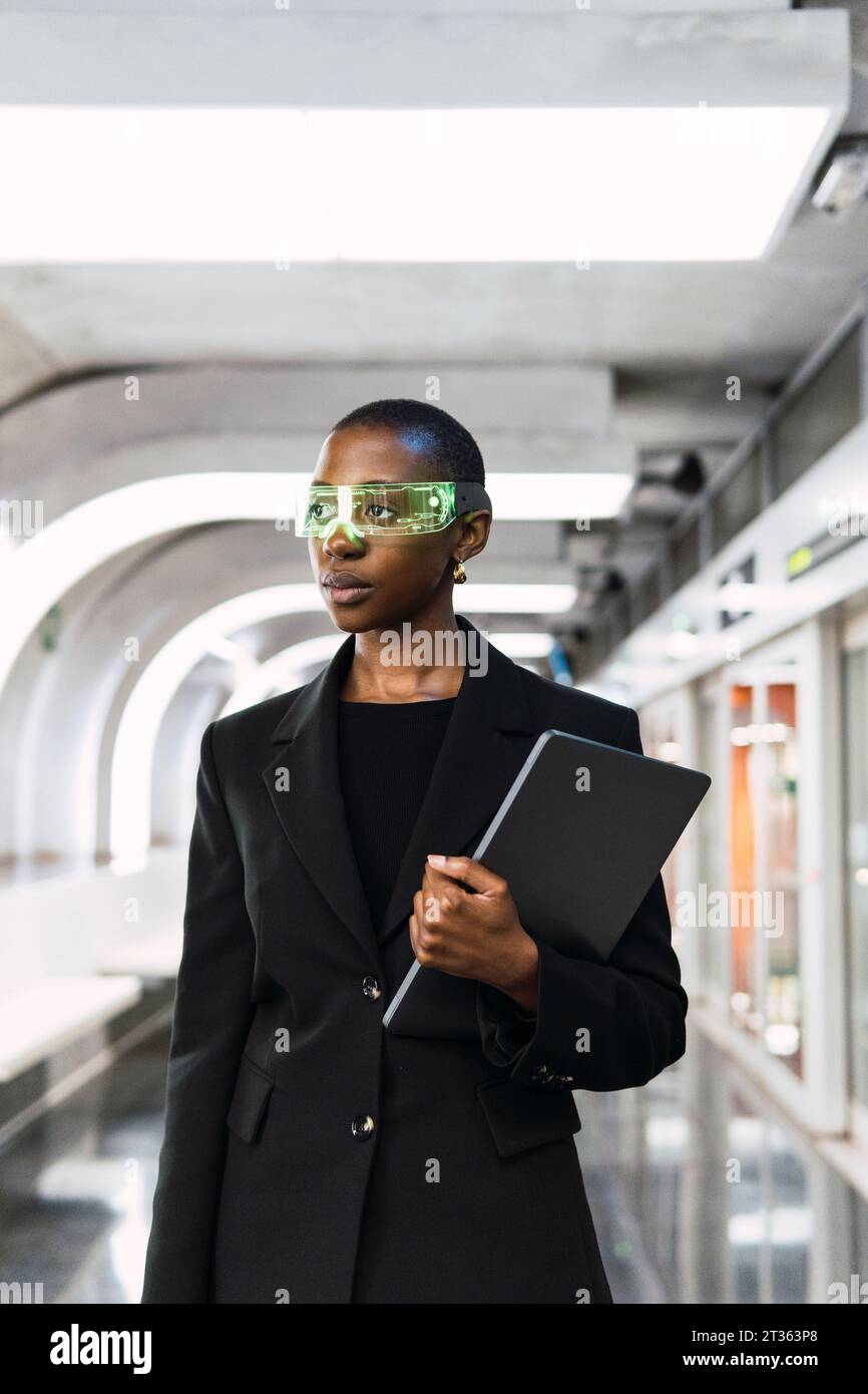 Donna contemplativa che indossa occhiali intelligenti e si trova in piedi con un computer portatile alla stazione della metropolitana Foto Stock