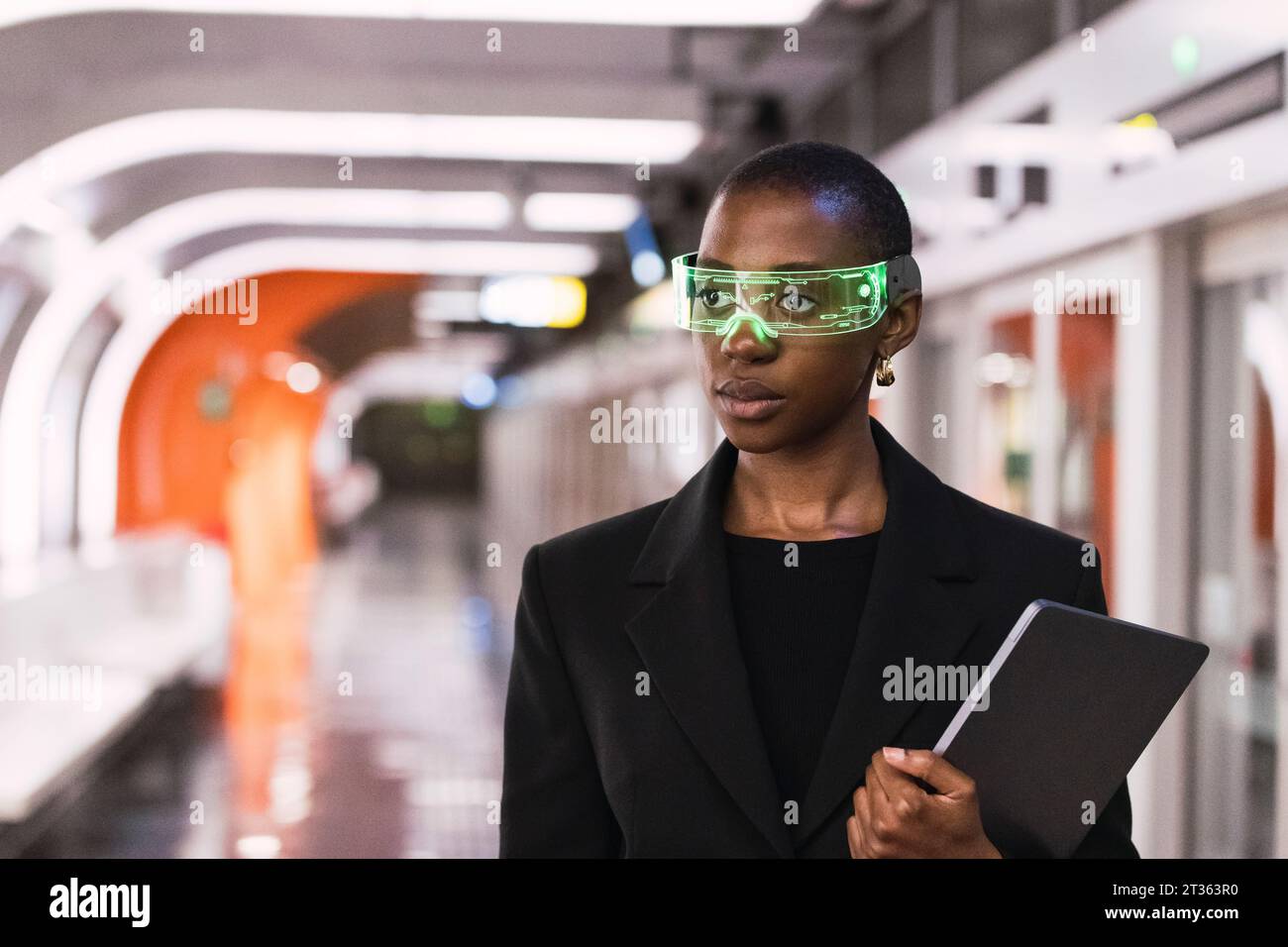 Donna contemplativa che indossa occhiali intelligenti e si trova in piedi con un computer portatile alla stazione della metropolitana Foto Stock