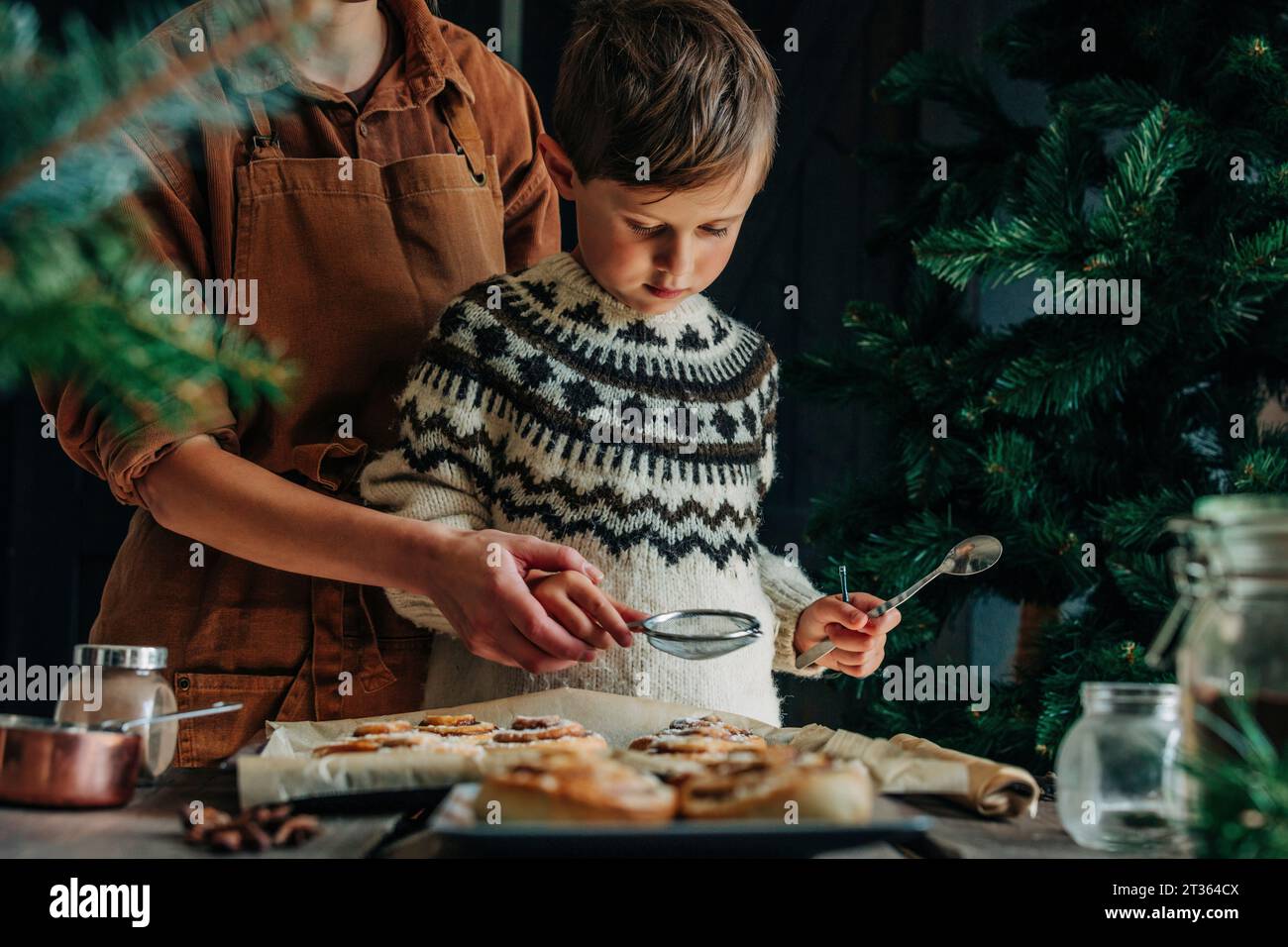 Madre e figlio spolverano zucchero sui panini alla cannella a tavola Foto Stock
