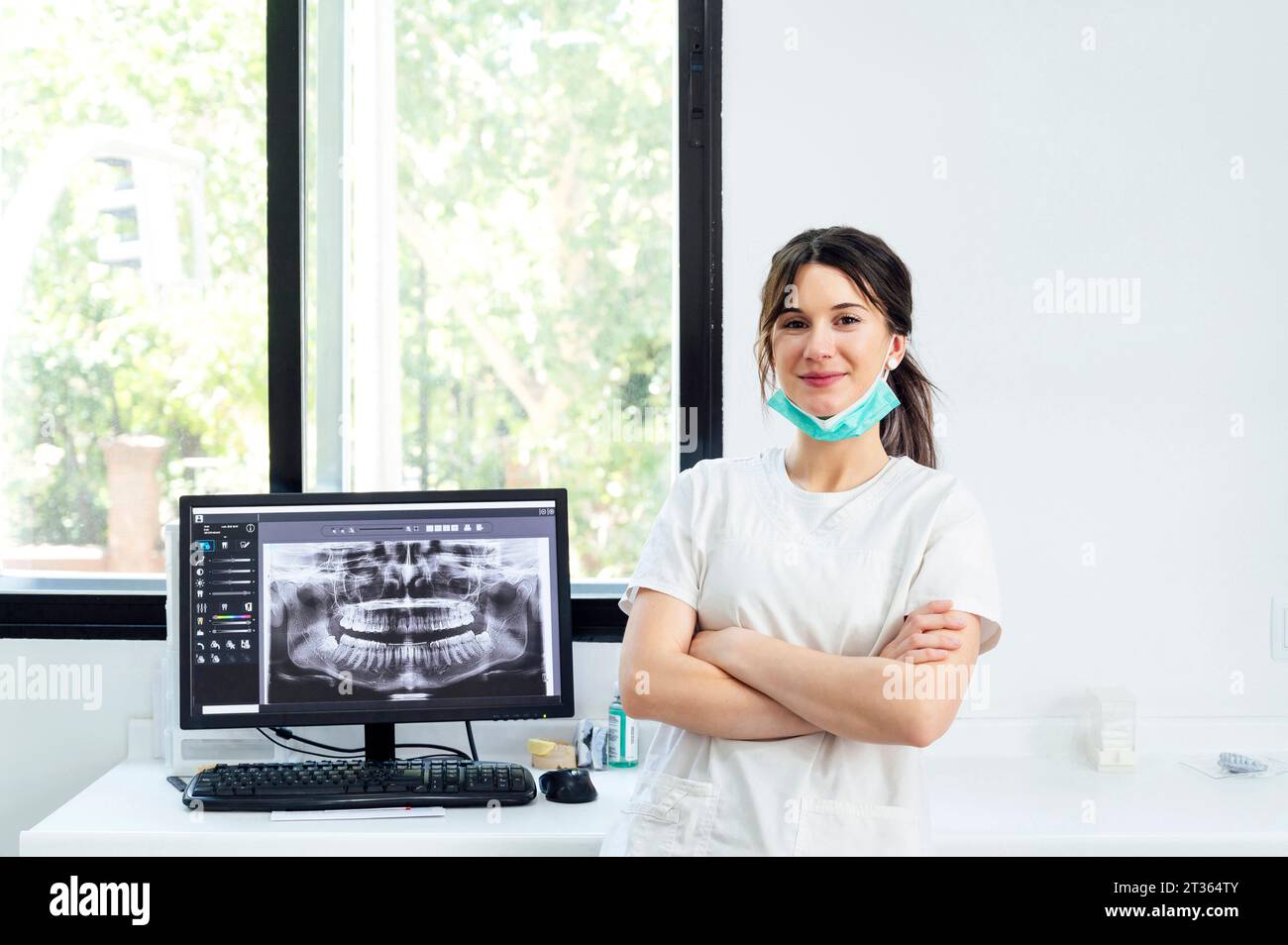 Dentista sorridente con le braccia incrociate in piedi vicino all'immagine radiografica sullo schermo del computer Foto Stock