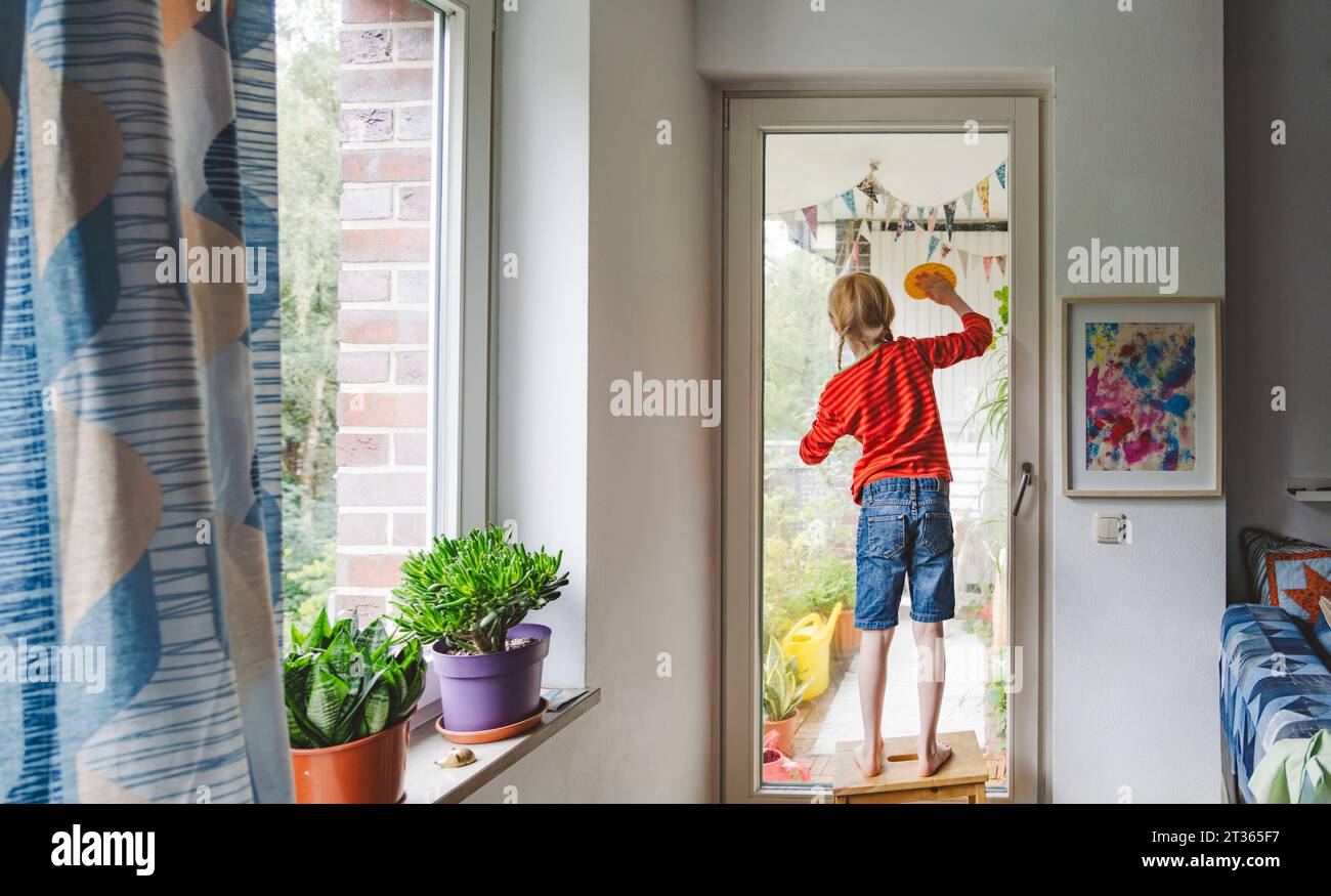 Ragazza che pulisce la porta del balcone in piedi sullo sgabello a casa Foto Stock