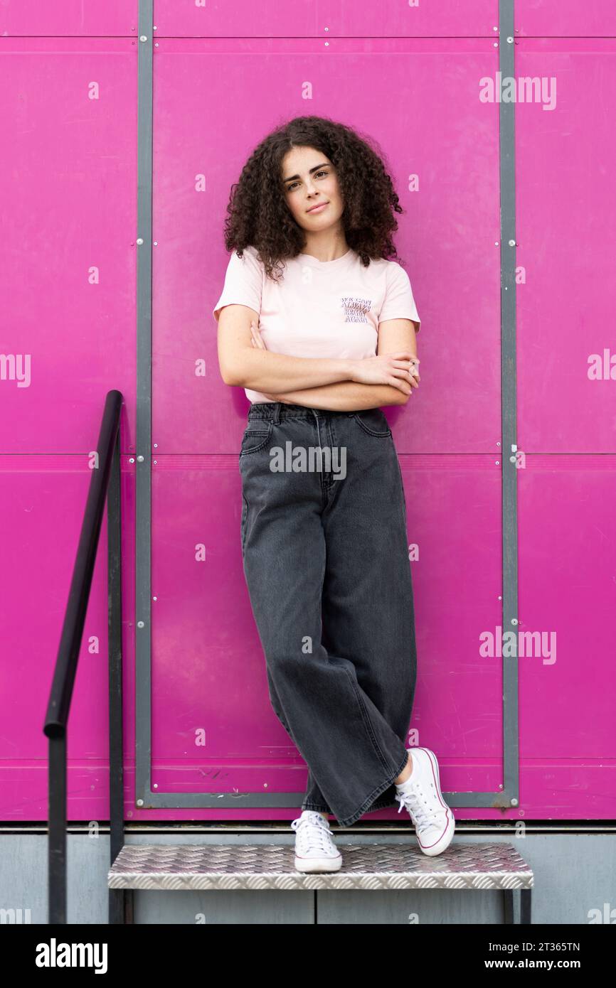 Giovane donna sorridente in piedi con le braccia incrociate davanti alla porta rosa Foto Stock