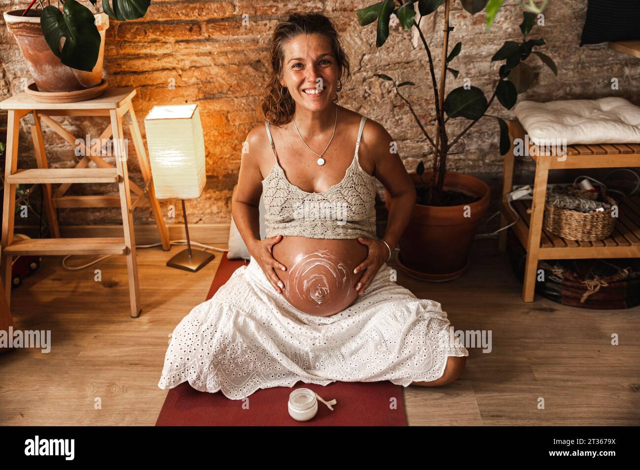 Donna incinta sorridente che applica idratante sull'addome a casa Foto Stock