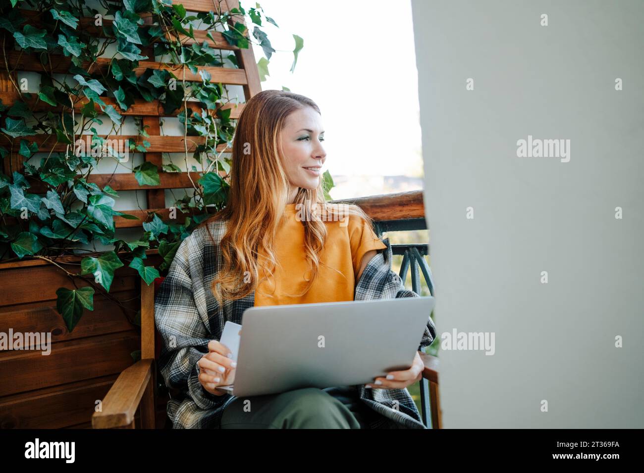 Donna sorridente con la rossa seduta con carta di credito e con il computer portatile nel balcone Foto Stock