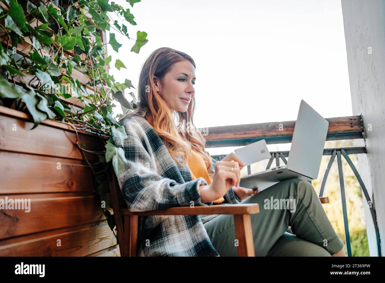 Donna sorridente con la rossa seduta con carta di credito e con il computer portatile nel balcone Foto Stock