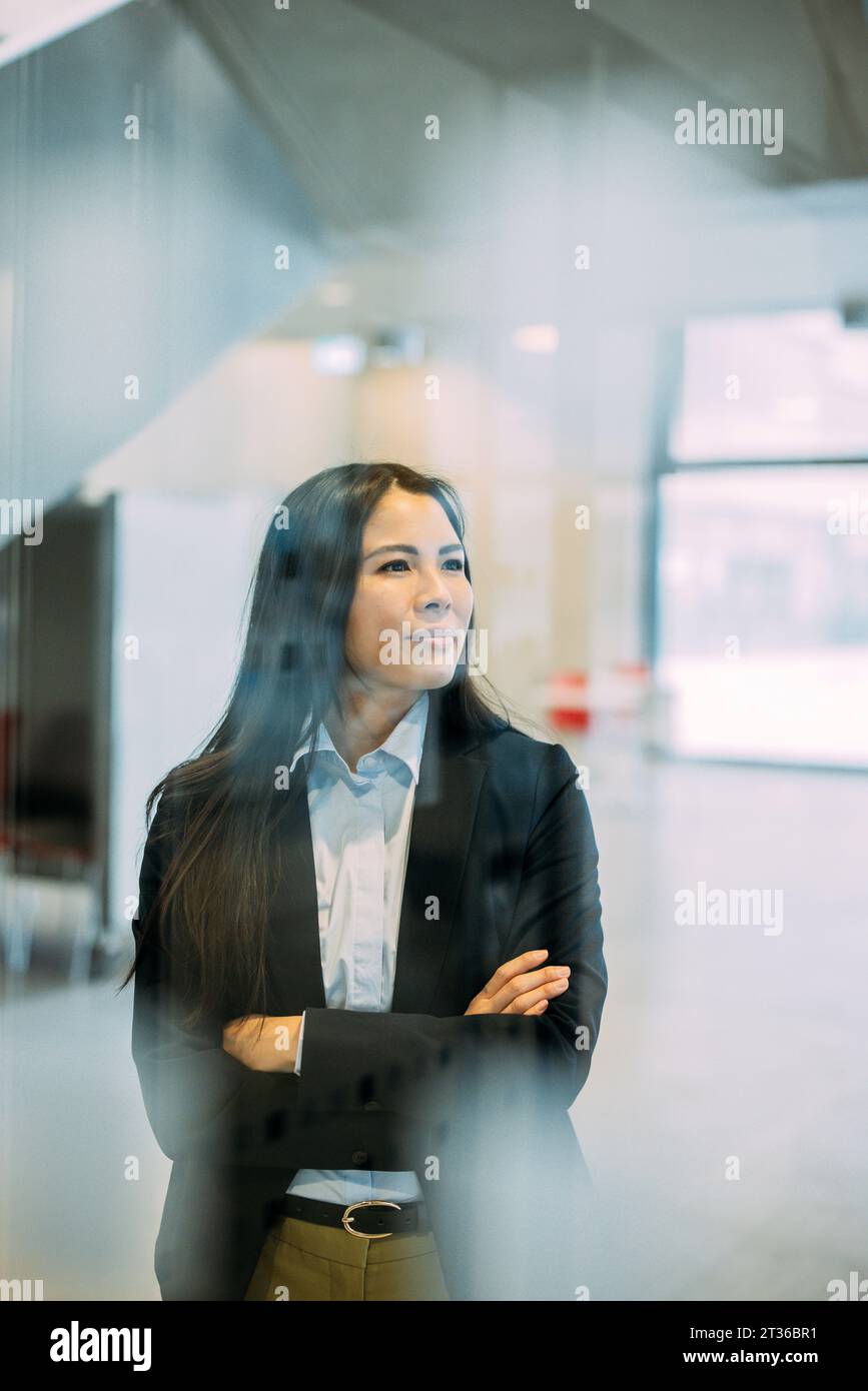 Donna d'affari in piedi con le braccia incrociate vista attraverso il vetro sul posto di lavoro Foto Stock