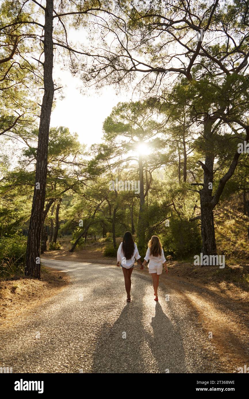 Ragazza adolescente che si tiene per mano con la madre che cammina su strada nella foresta al tramonto Foto Stock