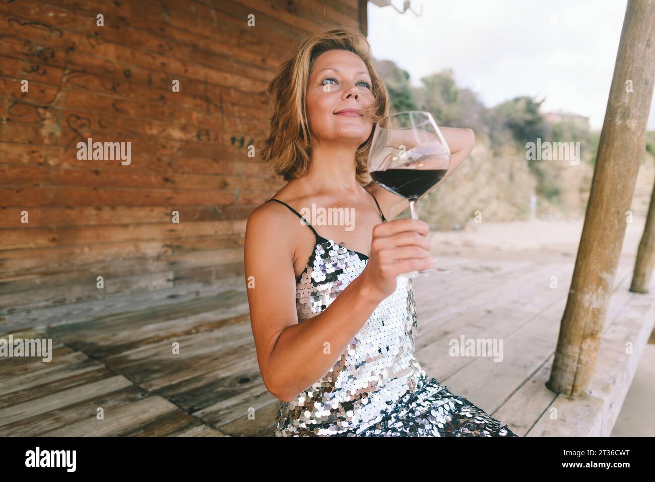 Donna sorridente che si gode un bicchiere di vino seduto su una veranda di legno Foto Stock