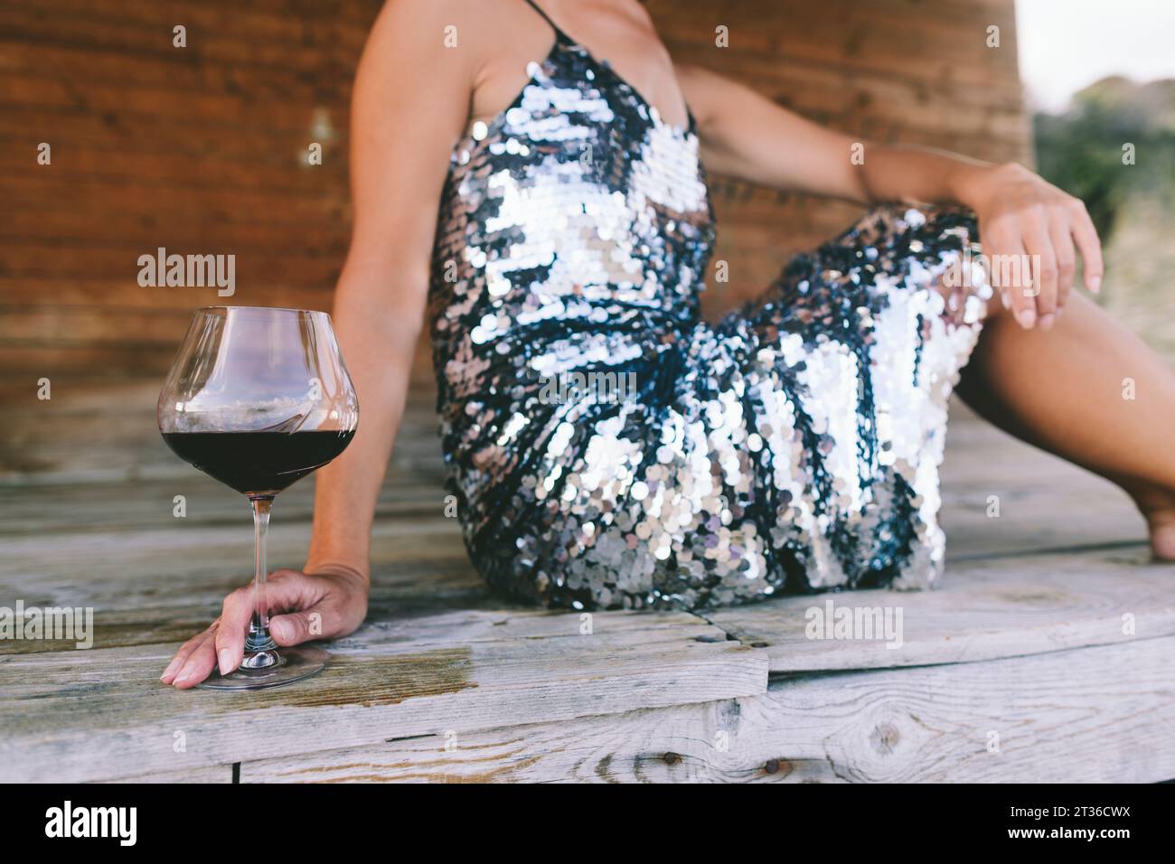 Donna che indossa un abito di paillettes seduta con wineglass su una veranda di legno Foto Stock