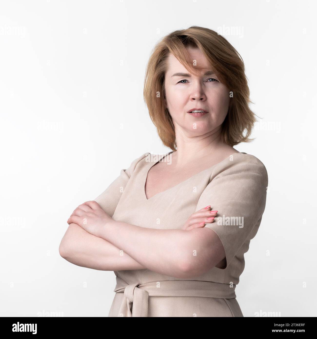 Ritratto di donna seria di 49 anni in abito beige con maniche corte e cintura allacciata davanti guardando con attenzione la fotocamera, con le braccia incrociate sul petto Foto Stock