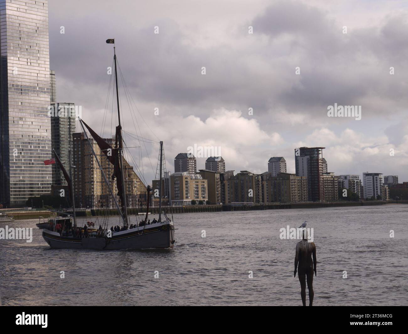 Limehouse, Londra, Regno Unito - 21 ottobre 2023: Antony Gormley's Another Time, con un gabbiano arroccato sulla testa, guarda passare la chiatta a vela del Tamigi. Foto Stock