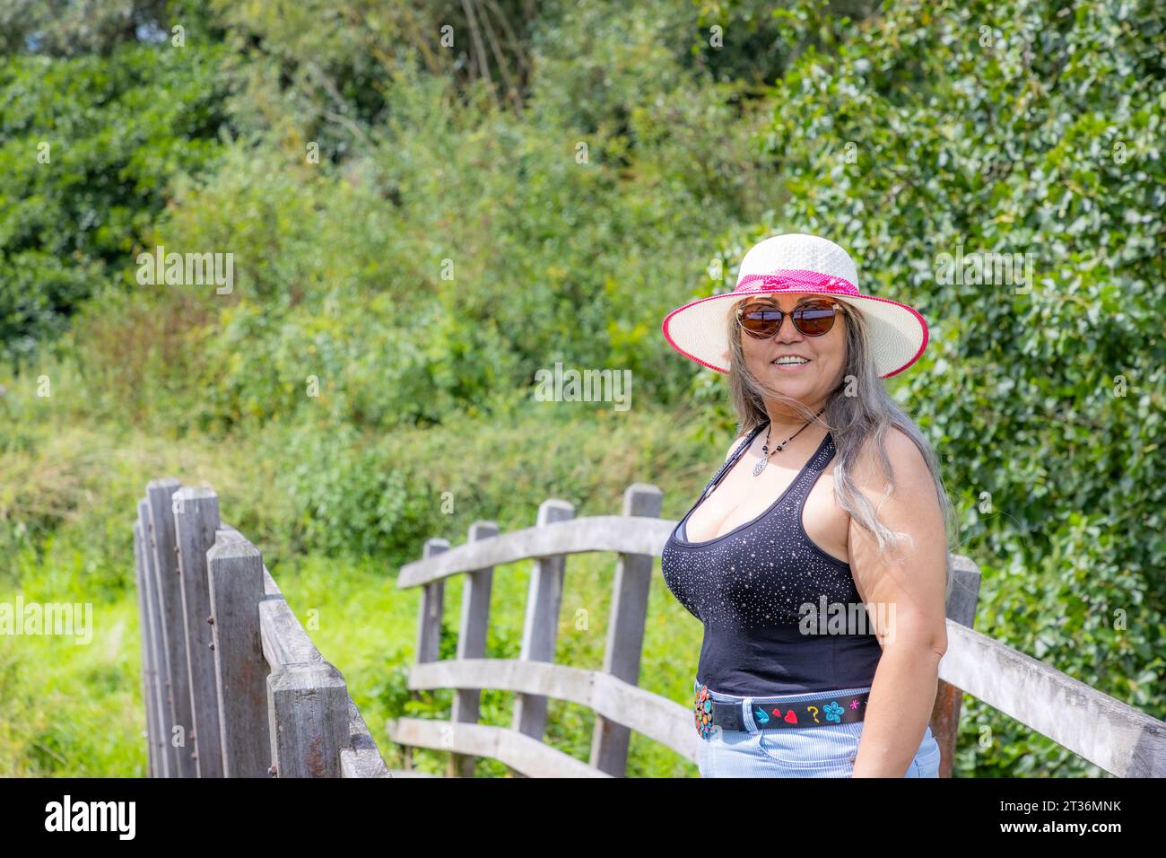 Chubby donna anziana adulta in piedi su un ponte di legno, che cerca la macchina fotografica, abiti casual, camicetta con corda, pantaloncini corti, cappello e occhiali da sole per godersi la giornata di sole Foto Stock