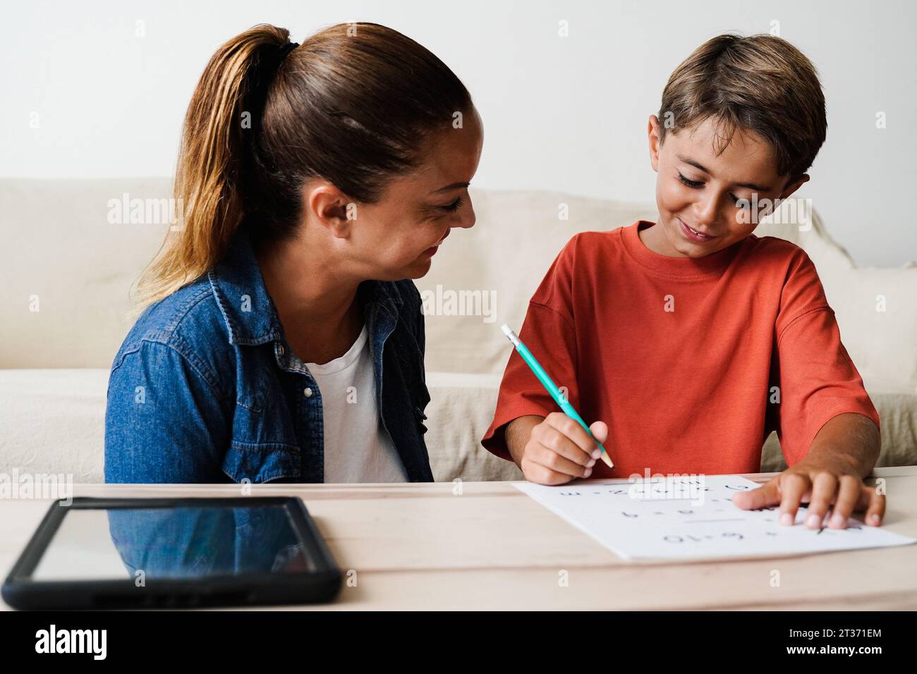 Madre e figlio latini che fanno i compiti per la scuola all'interno del salotto - i genitori aiutano il bambino con l'istruzione scolastica Foto Stock
