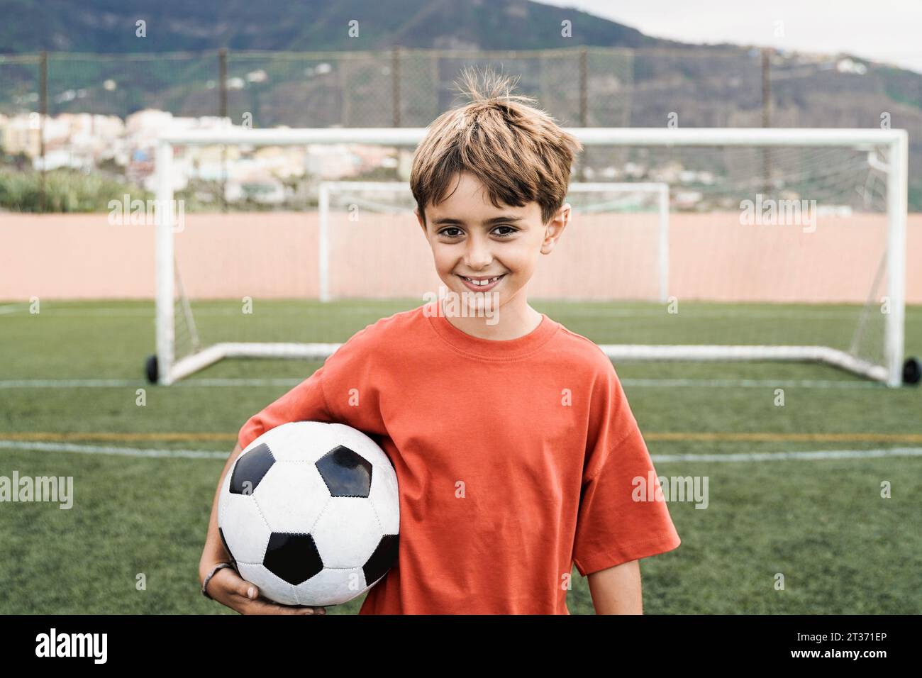 Bambino bambino bambino giocatore di calcio che tiene palla con campo di calcio sullo sfondo - bambini sport stile di vita Foto Stock