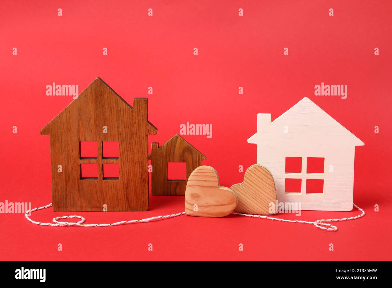 Cuori in legno e cordoncino decorativo tra i modelli di casa su sfondo rosso che simboleggia la connessione in relazione a lunga distanza Foto Stock