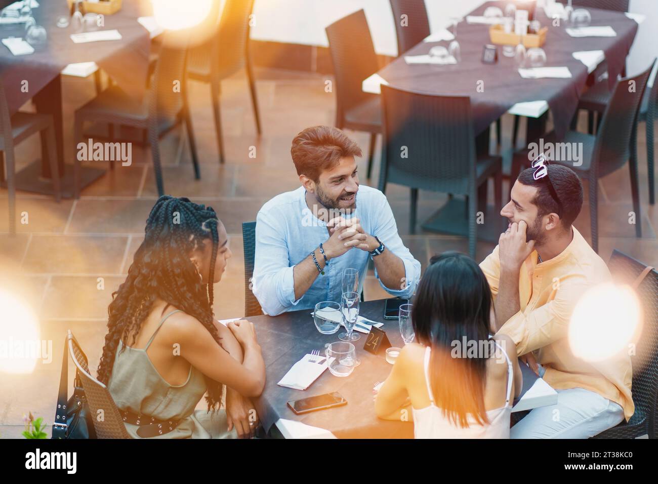 Un gruppo multiculturale di amici si riunisce in un ambiente all'aperto, condividendo risate e conversazioni. Seduti a un tavolo, si godono un pasto e l'un l'altro Foto Stock