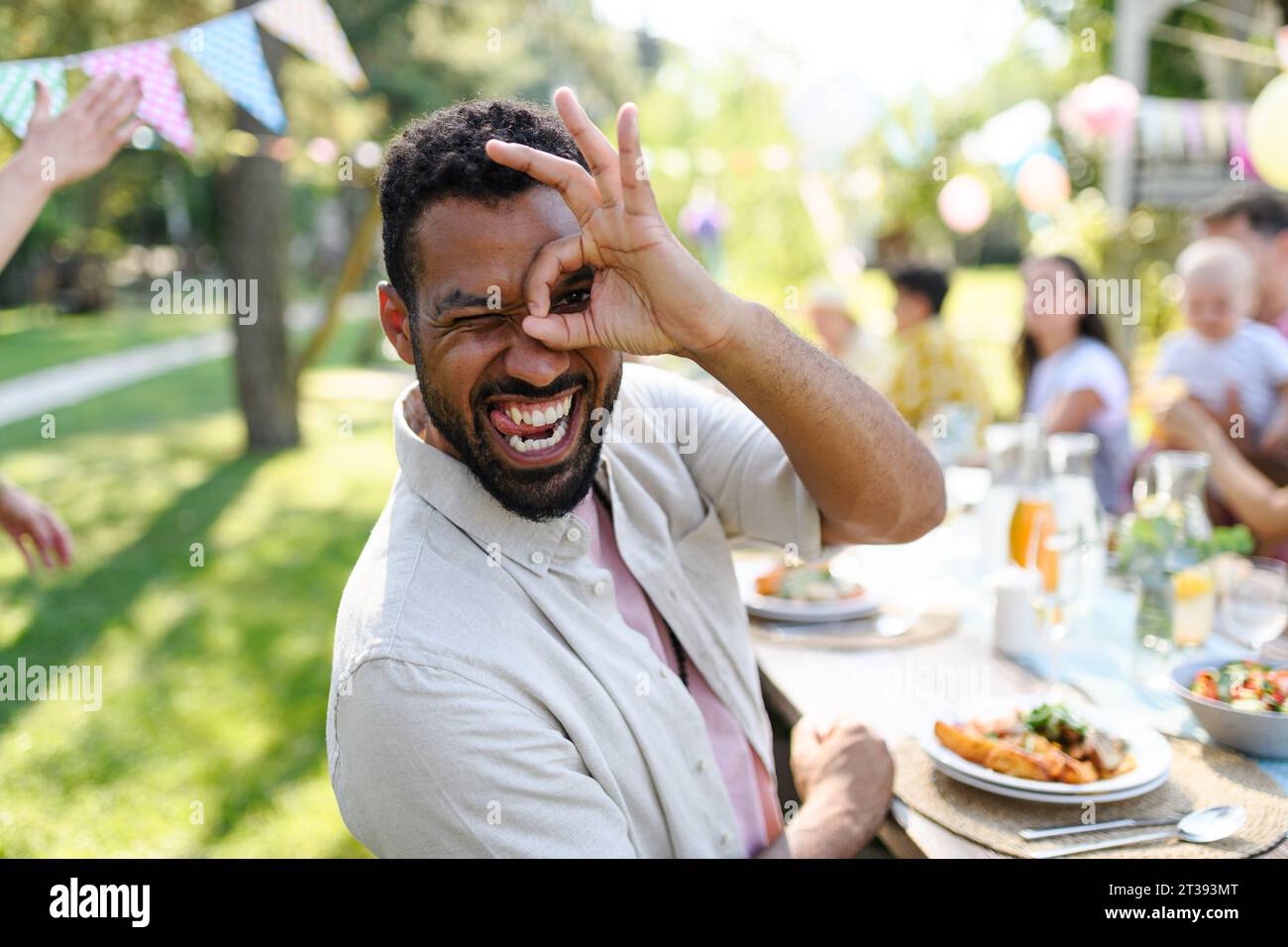 Bell'uomo che fa facce divertenti, seduto al tavolo della festa durante una festa estiva in giardino con la famiglia e gli amici. Foto Stock