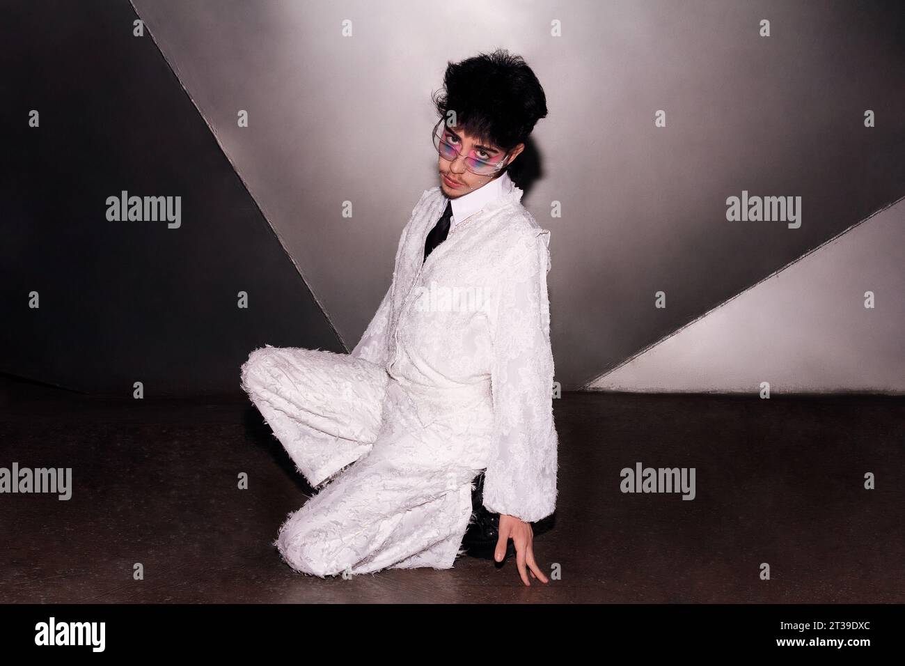Persona alla moda con un trucco sorprendente, seduta su un pavimento di cemento con un'elegante tuta bianca, che offre uno sguardo forte verso l'agitazione della fotocamera Foto Stock