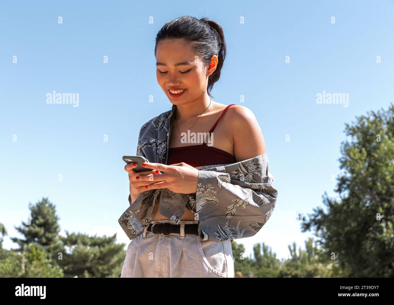 Giovane donna sorridente che usa il suo smartphone all'aperto in una giornata di sole, vestita con un abito elegante. Foto Stock