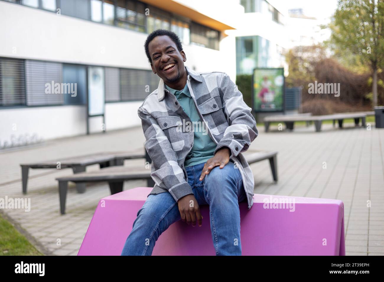 Ridere il giovane uomo afroamericano con gli occhi chiusi seduto su una panchina di cemento vicino al marciapiede di fronte a alberi verdi sfuocati alla luce del giorno Foto Stock