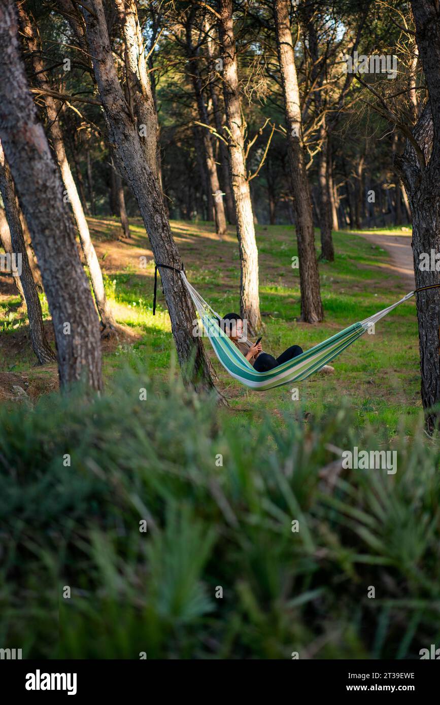 Vista laterale di una donna negli occhiali che guarda lo schermo del telefono cellulare mentre riposa su un'amaca a strisce legata ai tronchi degli alberi della foresta andalusa in giorno Foto Stock