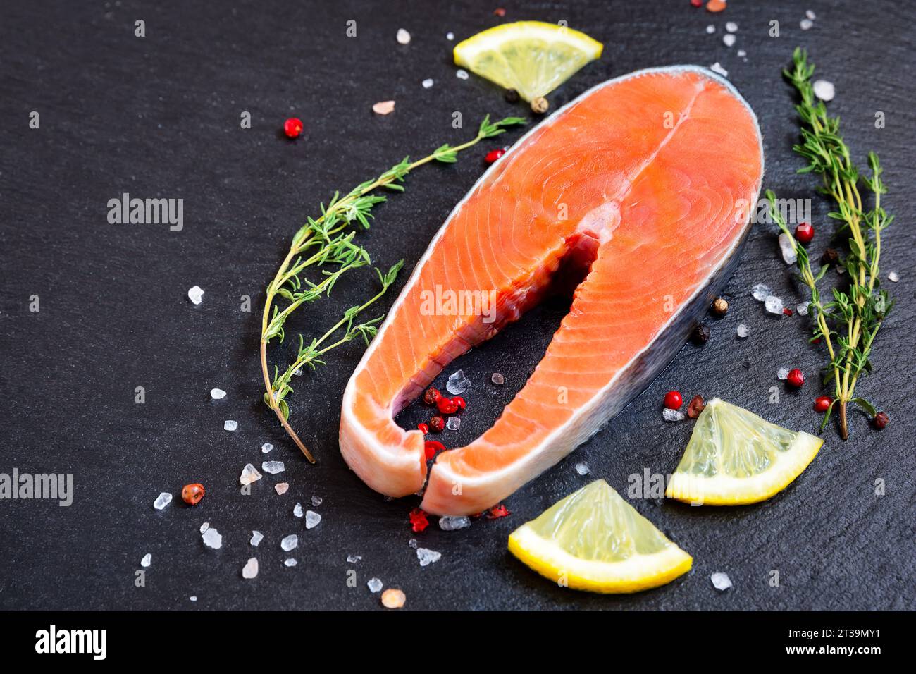 Bistecca di pesce fresco crudo con ingredienti da cucina, erbe aromatiche e limone su sfondo nero, vista dall'alto. Bistecca di salmone con timo e limone su tavola nera. Foto Stock