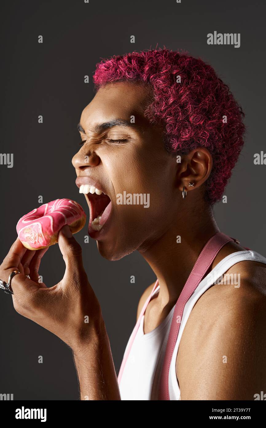 bellissimo uomo elegante con capelli rosa e accessori argentati che mangiano ciambella rosa, moda e stile Foto Stock