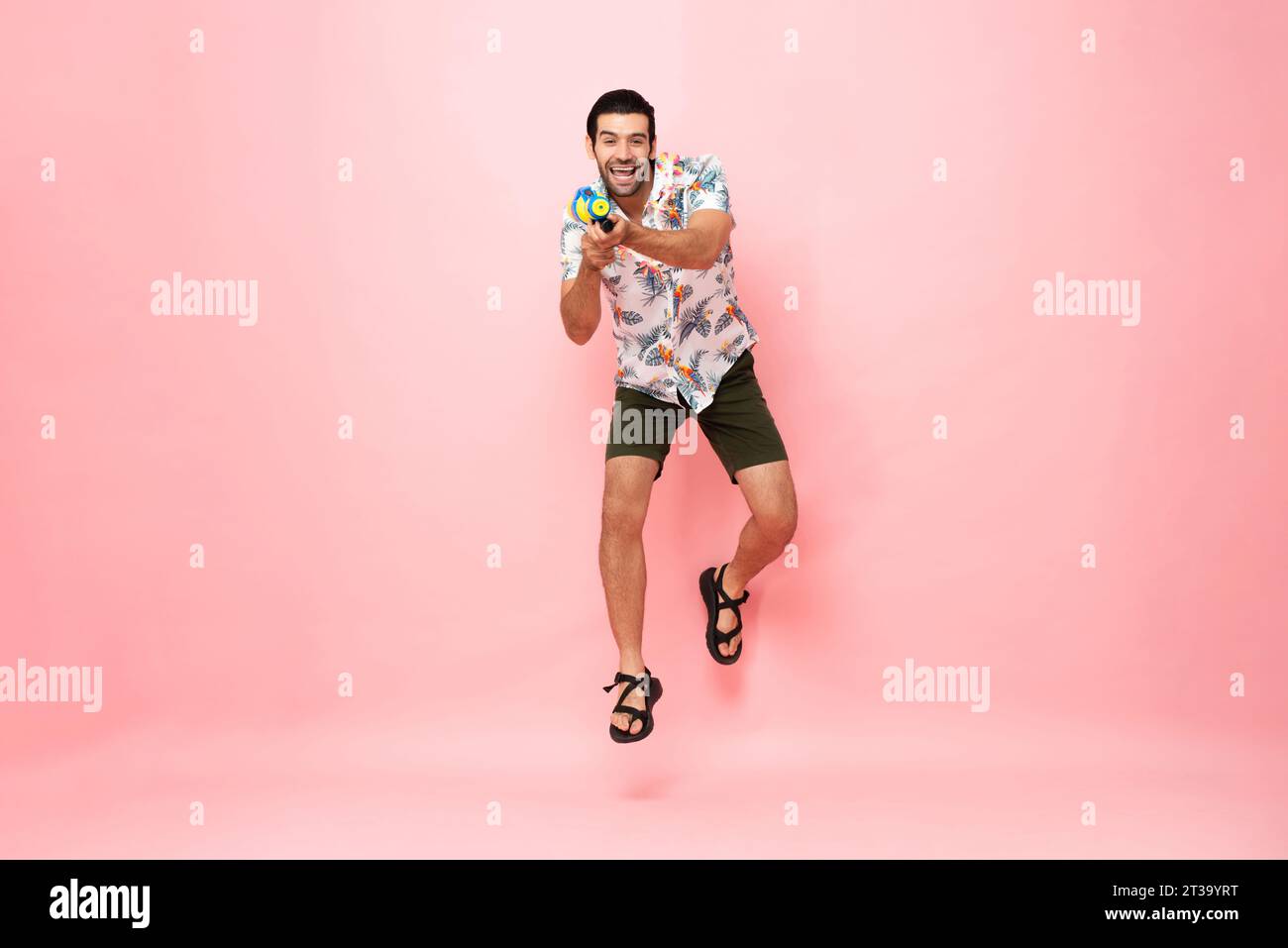 Bellissimo e sorridente turista caucasico che gioca con la pistola ad acqua e salta in uno sfondo rosa studio per il festival Songkran in Thailandia An Foto Stock
