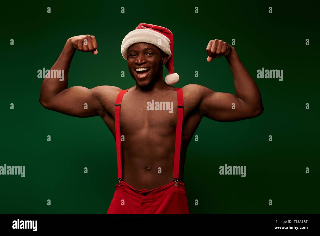 uomo afro-americano felice e forte con cappello e pantaloni natalizi che mostrano i muscoli su sfondo verde Foto Stock