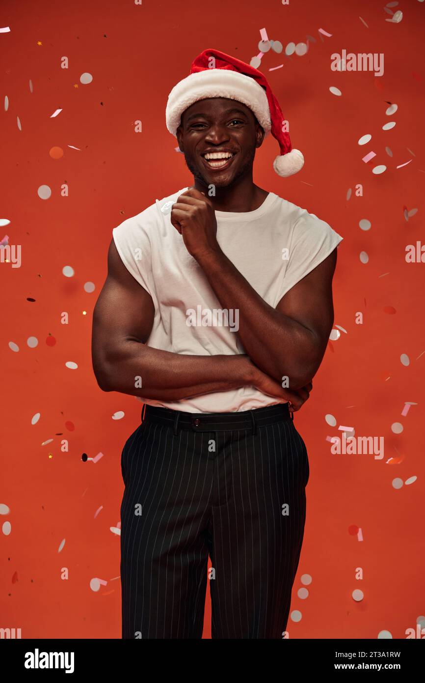 gioioso uomo afro-americano con cappello di natale che guarda la macchina fotografica su sfondo rosso con coriandoli cadenti Foto Stock