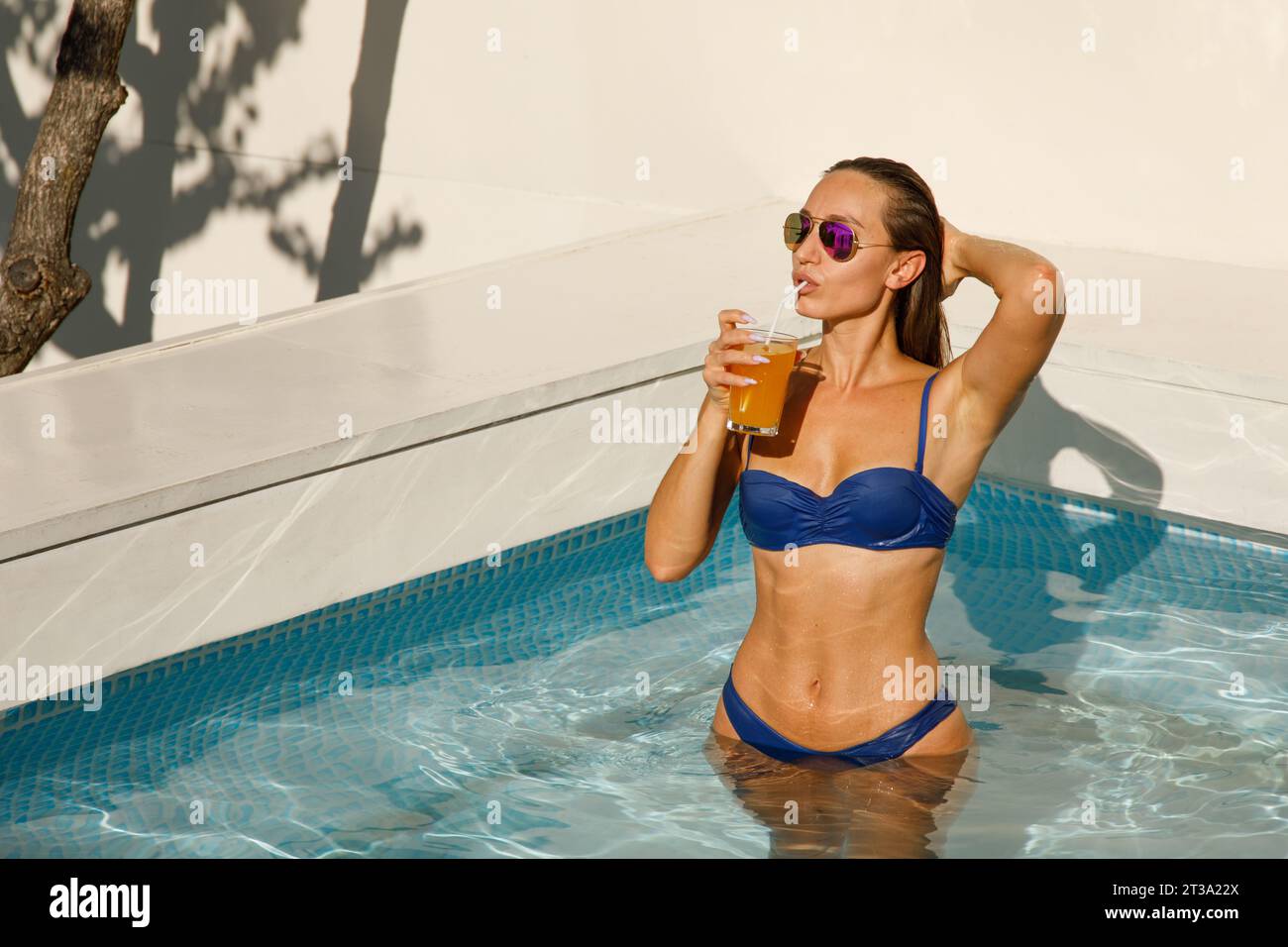 Elegante ritiro in piscina: Donna alla moda che sorseggia succo d'arancia con occhiali da sole Foto Stock