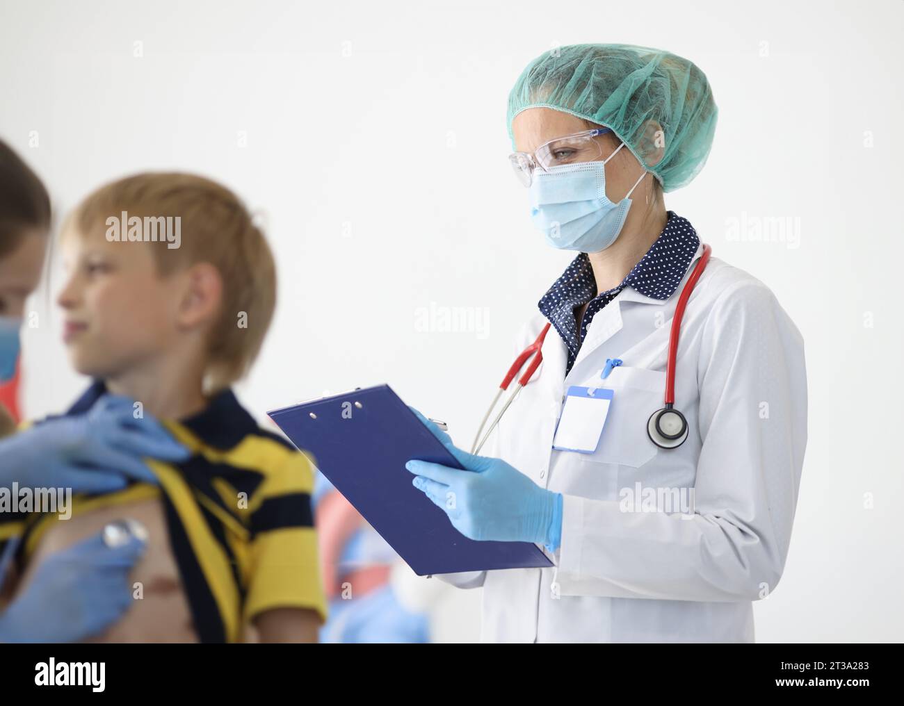 Medico in maschera medica protettiva scrive le letture sulla clipboard accanto al medico ascolta con stetoscopio al respiro del ragazzo. Malattia dell'infanzia prefilassi Foto Stock