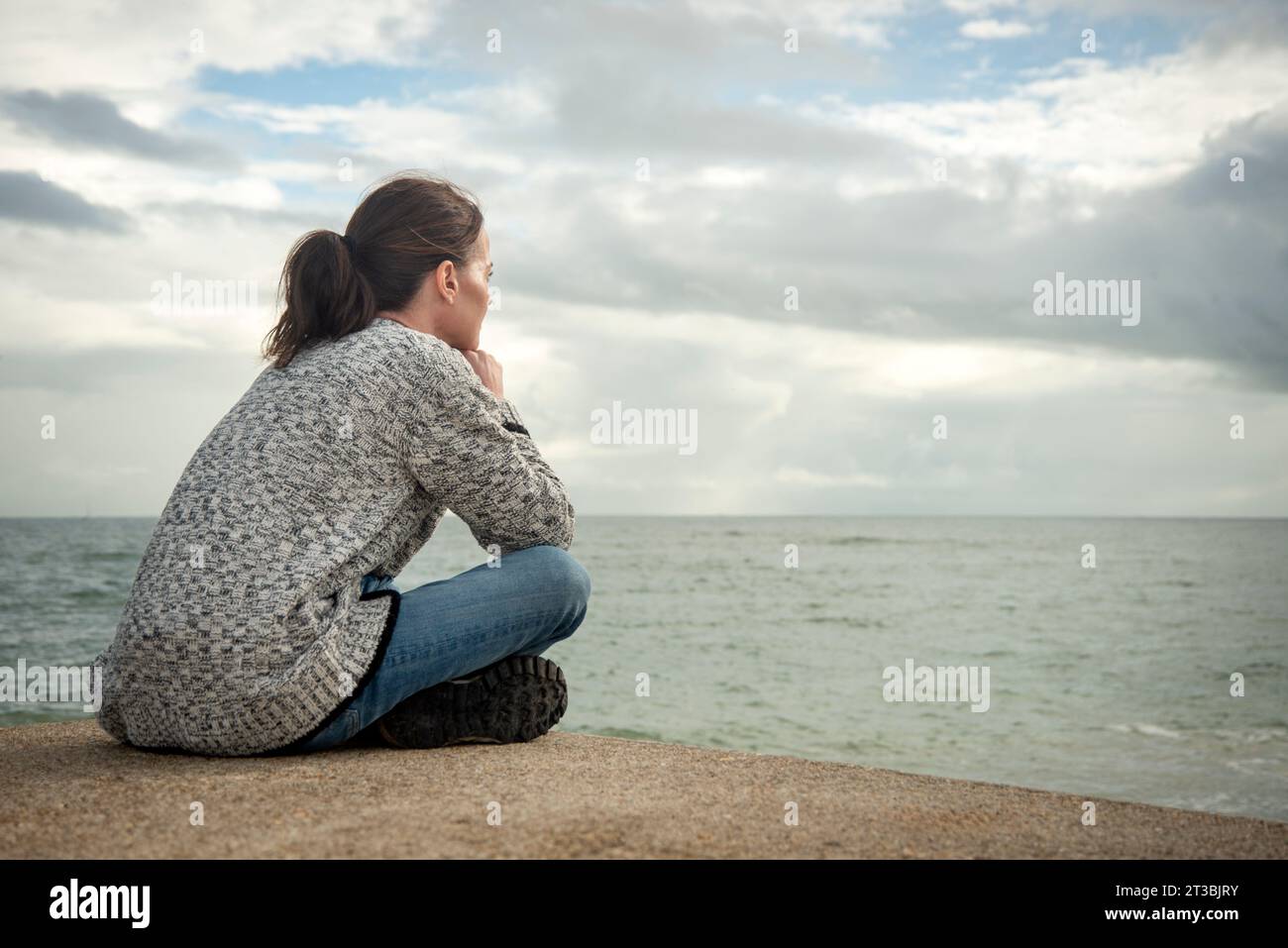 Donna seduta da sola che guarda il mare nel profondo del pensiero, vista posteriore, autunno. Foto Stock