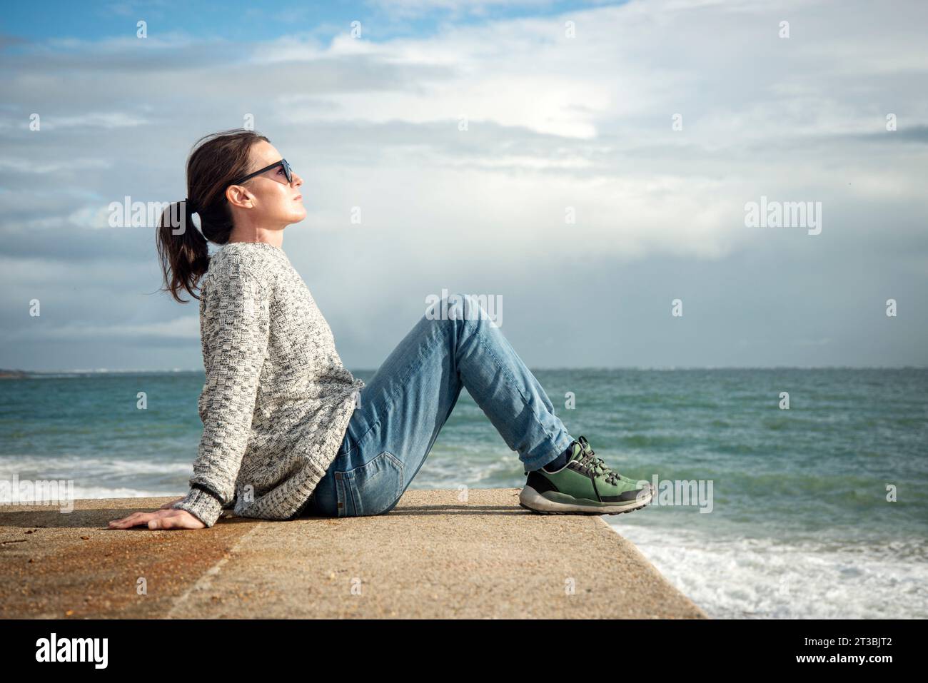 donna che indossa un maglione e occhiali da sole che si rilassa e si gode il sole invernale e autunnale in riva al mare Foto Stock