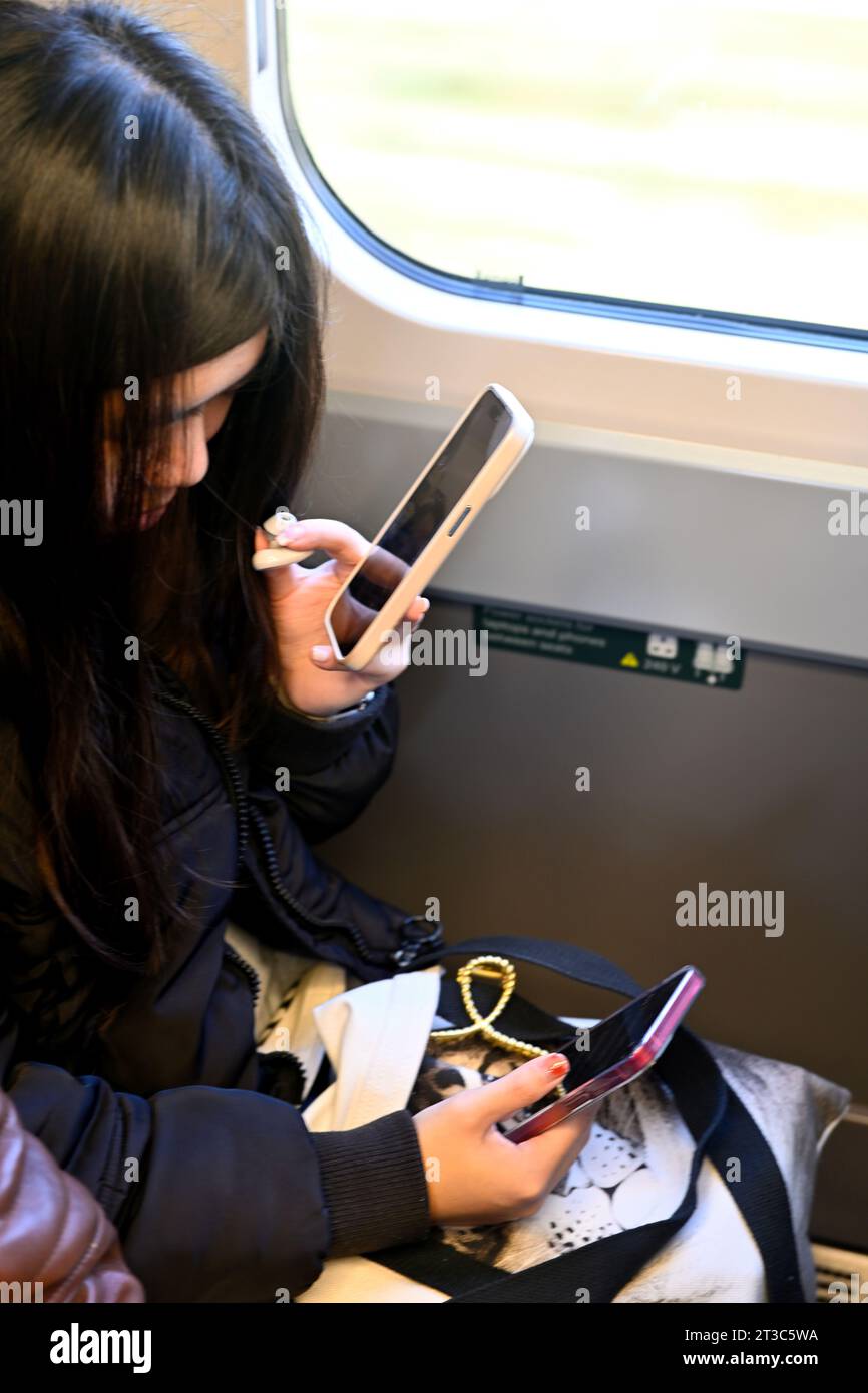 Giovane donna adulta che utilizza due telefoni cellulari contemporaneamente quando viaggia in treno, una voce una digitazione, Regno Unito Foto Stock