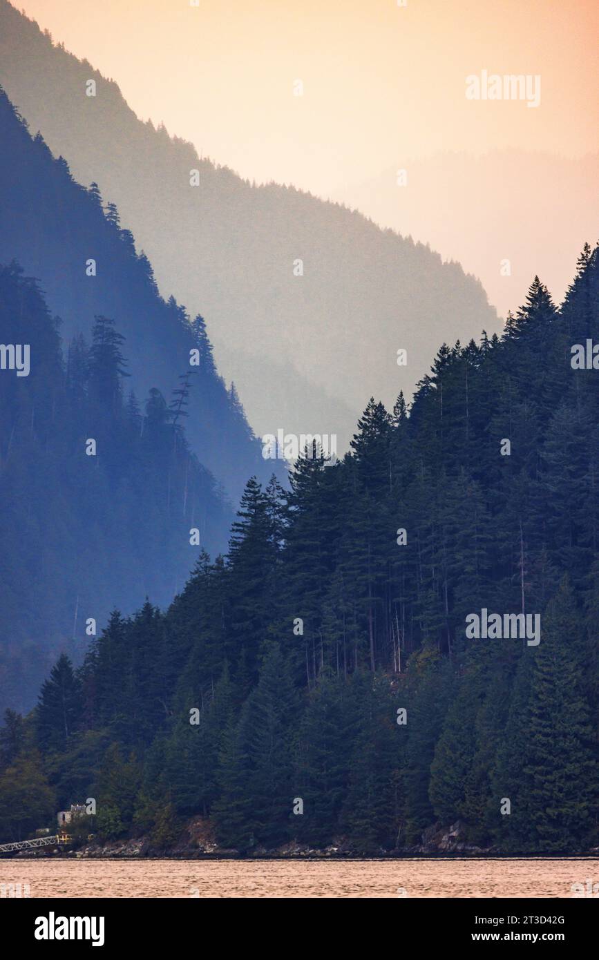 Le montagne nel remoto braccio Indiano svaniscono nel cielo rese arancioni dal fumo di fuoco nella Columbia Britannica, Canada. Vista del paesaggio da una barca sul fiordo. Foto Stock